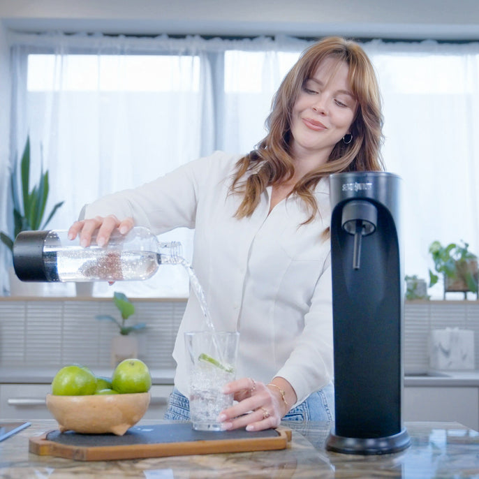 Person pouring sparkling water from a Soda Sense bottle into a glass with lime slices in a bright, modern kitchen. A black Soda Sense soda maker and a wooden bowl of fresh limes are on the counter.