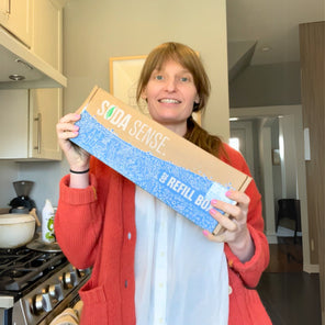 A customer smiling while holding a Soda Sense CO2 canister refill box, ready to be mailed for exchange.